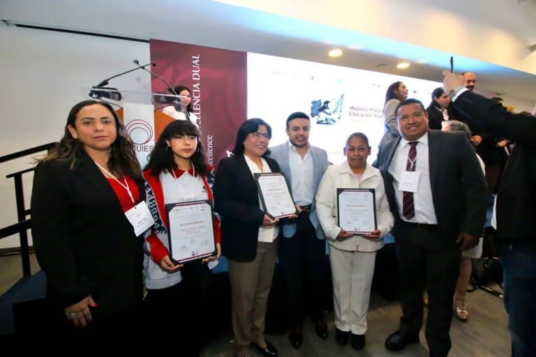 Grupo de personas en una ceremonia de reconocimiento, algunos sosteniendo certificados, en un evento formal en un auditorio con pantalla grande al fondo.