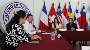 Panel de discusión con representantes de diferentes países en Tlaxcala, con banderas internacionales al fondo y participantes en la mesa de debate.