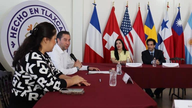 Panel de discusión con representantes de diferentes países en Tlaxcala, con banderas internacionales al fondo y participantes en la mesa de debate.