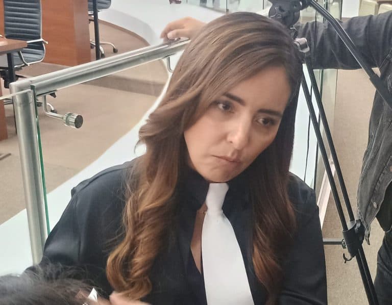 Asistente de prensa en una conferencia, mujer con cabello castaño y ondulado, vestida con traje negro y corbata blanca, en un entorno formal con equipo de grabación y micrófonos.