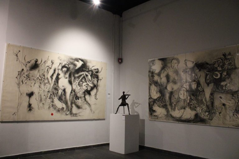 Dos grandes obras de arte en tonos negros y grises en una galería, con una escultura en el centro en exhibición.