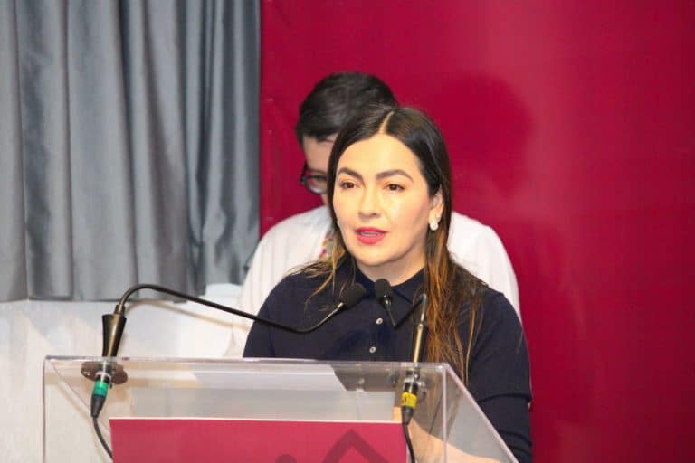 La mujer habla en un podio durante una conferencia o evento formal, con fondo rojo y cortinas grises a su lado.
