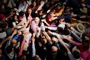 Joven mujer rodeada de muchas personas en un evento público, saludando y sonriendo, en un ambiente de interacción social y celebración.