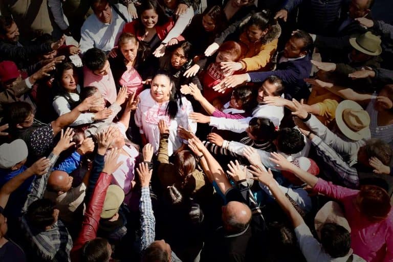 Joven mujer rodeada de muchas personas en un evento público, saludando y sonriendo, en un ambiente de interacción social y celebración.