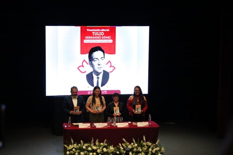 Cuatro personas en una mesa durante una presentación o evento en un escenario con pantalla grande y decoraciones florales en primer plano.