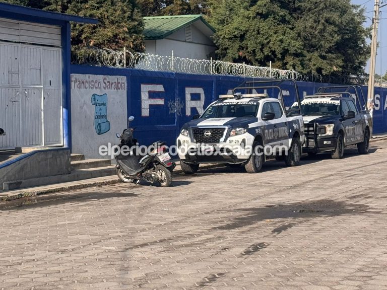 Vehículos de policía y una motocicleta en la calle frente a una barda con alambre de púas en Tlaxcala, México, en una escena que sugiere presencia policial o un incidente en la zona.