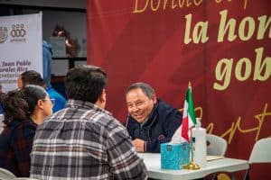 Asesor político conversando con ciudadanos en un evento público en Tlaxcala, México, con fondo rojo y bandera mexicana en la mesa.