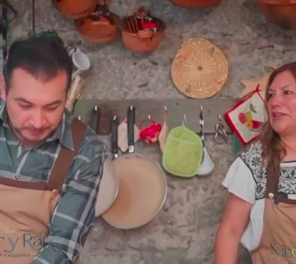 Cocinero y asistente en una cocina tradicional con utensilios colgados en la pared de piedra.