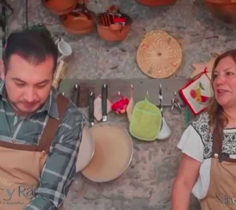 Cocinero y asistente en una cocina tradicional con utensilios colgados en la pared de piedra.