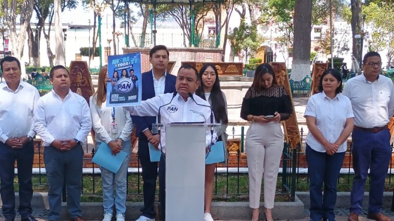Manifestante en un mitin político en Tlaxcala, con un cartel del PAN y un micrófono, rodeado de varias personas en un espacio público con árboles y bancos, durante el día.