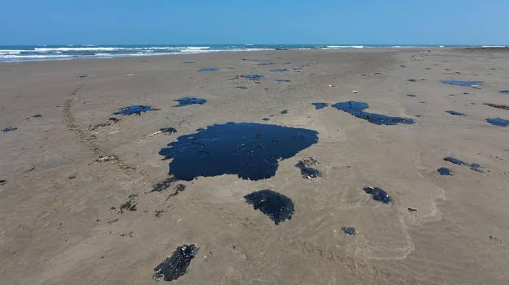 Aceite derramado en la playa, formando manchas negras sobre la arena en un día soleado con el mar de fondo.