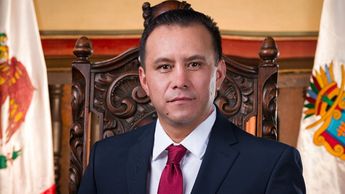 Elegante hombre en traje formal sentado en una silla de madera con banderas de fondo, en un entorno oficial y gubernamental en Tlaxcala, México.