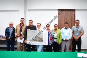 Grupo de ocho personas en una reunión formal, una de ellas sostiene un mapa o plano grande, en un entorno interior con paredes blancas y puerta de madera.
