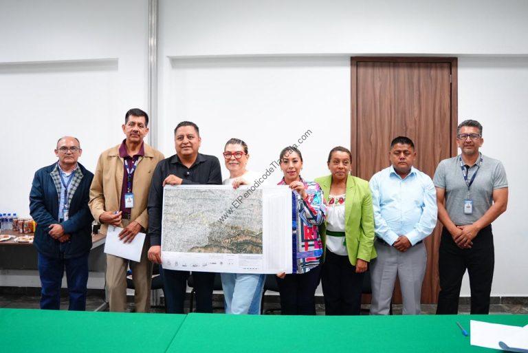 Grupo de ocho personas en una reunión formal, una de ellas sostiene un mapa o plano grande, en un entorno interior con paredes blancas y puerta de madera.