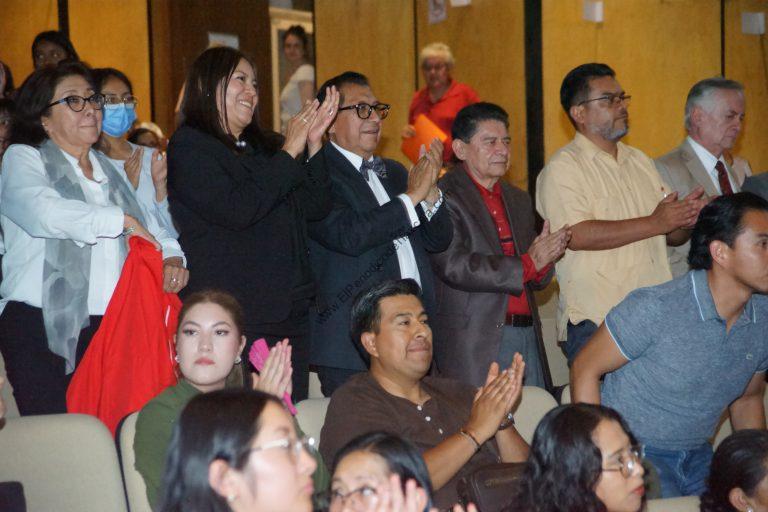 Grupo de personas en un evento formal, algunos aplaudiendo y otros observando, en un ambiente interior con fondo de madera y varias personas en el público.