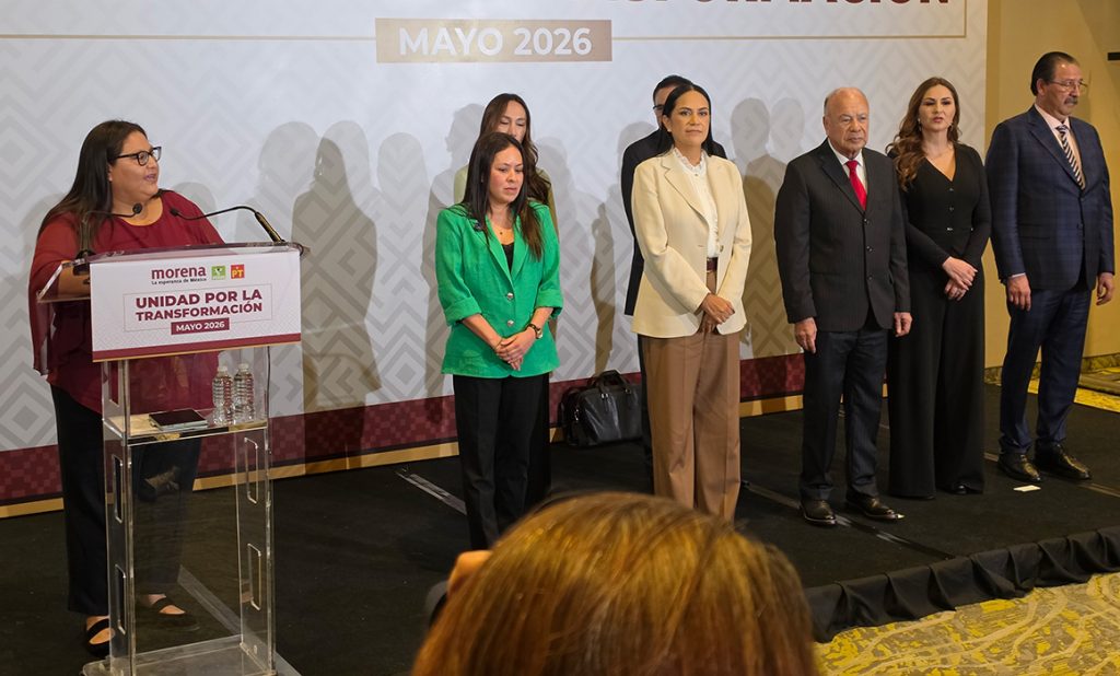 Grupo de seis personas en un escenario durante un evento político en mayo de 2026, con un podio y un cartel de Morena en el fondo.
