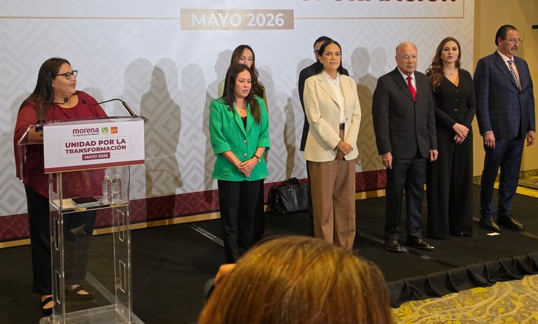Grupo de seis personas en un escenario durante un evento político en mayo de 2026, con un podio y un cartel de Morena en el fondo.