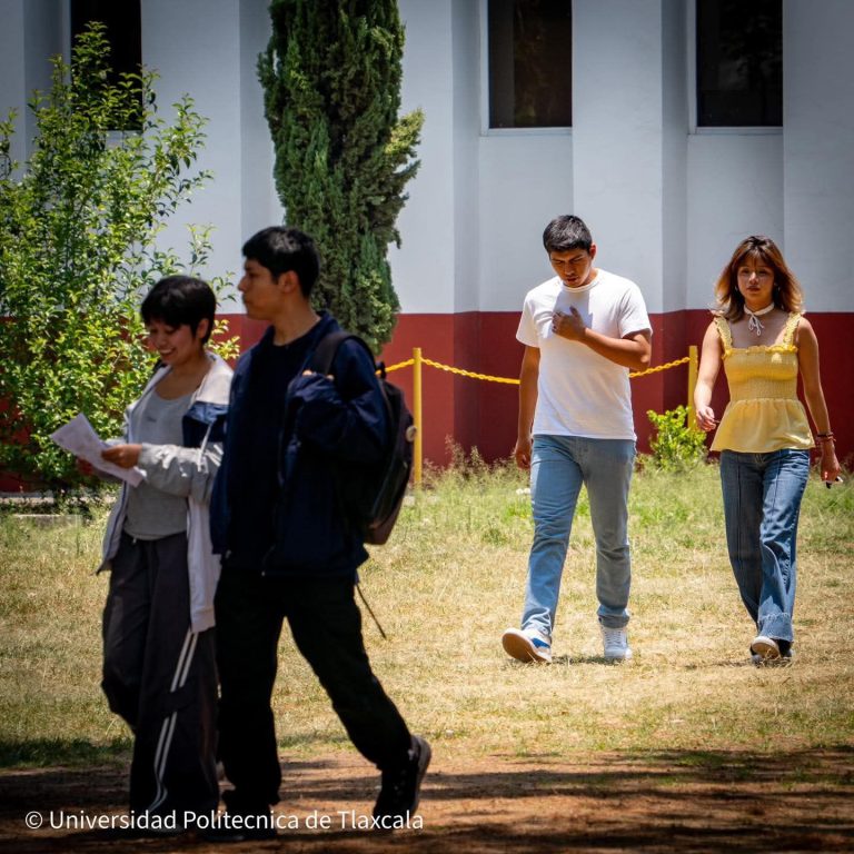 Estudiante universitario caminando en el campus de la Universidad Politécnica de Tlaxcala, con otros estudiantes en el fondo, en un día soleado y con vegetación alrededor.