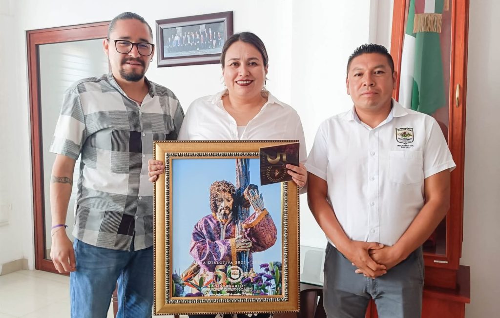 Tres personas en una oficina, una de ellas sostiene un retrato de un artista con un micrófono, en un entorno formal con bandera mexicana y cuadro en la pared.
