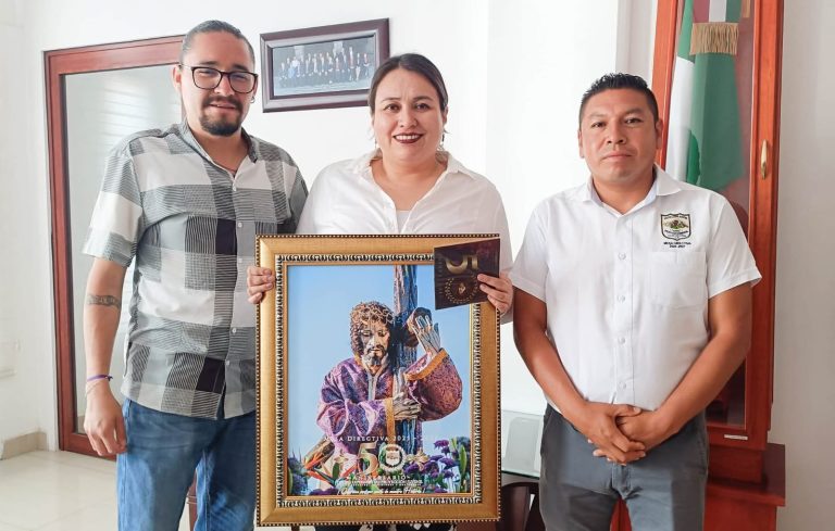 Tres personas en una oficina, una de ellas sostiene un retrato de un artista con un micrófono, en un entorno formal con bandera mexicana y cuadro en la pared.