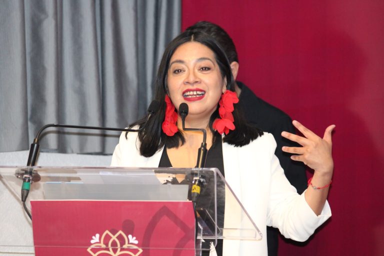 Mujer hablando en un evento formal, con cabello oscuro y vestido blanco, en un escenario con micrófono y fondo gris y rojo.