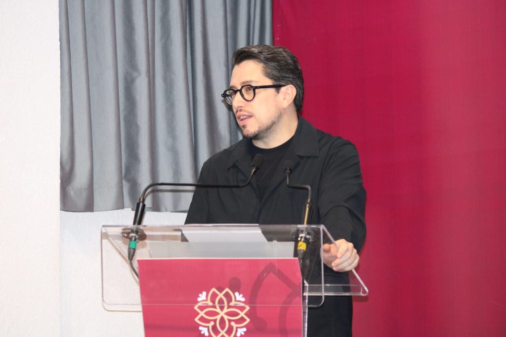 Hombre con gafas y barba hablando en un podio transparente en un evento formal con fondo de cortinas gris y rosa.