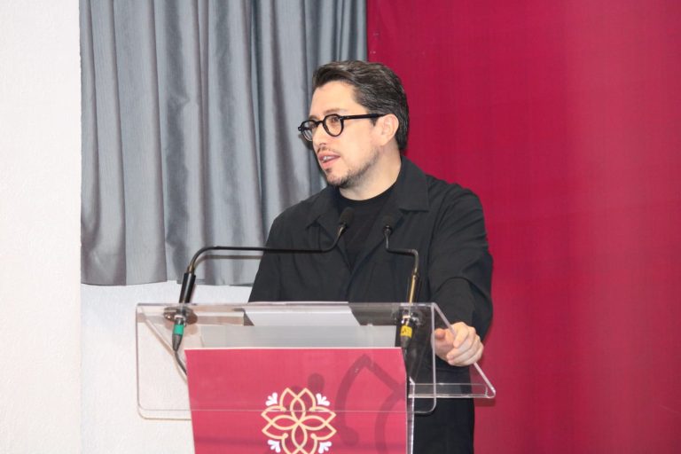 Hombre con gafas y barba hablando en un podio transparente en un evento formal con fondo de cortinas gris y rosa.