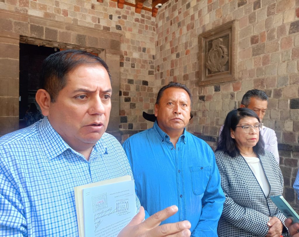 Hombre con camisa azul y expresión seria, sosteniendo documentos, en un entorno de ladrillos y piedra, acompañado por otras personas en una reunión o evento oficial.
