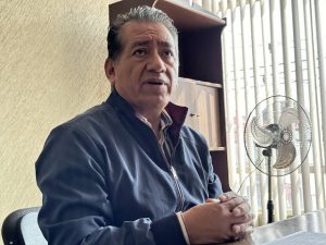 Hombre mayor sentado en una oficina, con expresión pensativa, vestido con chaqueta azul y camisa a rayas, en un ambiente con cortinas y ventilador en el fondo.