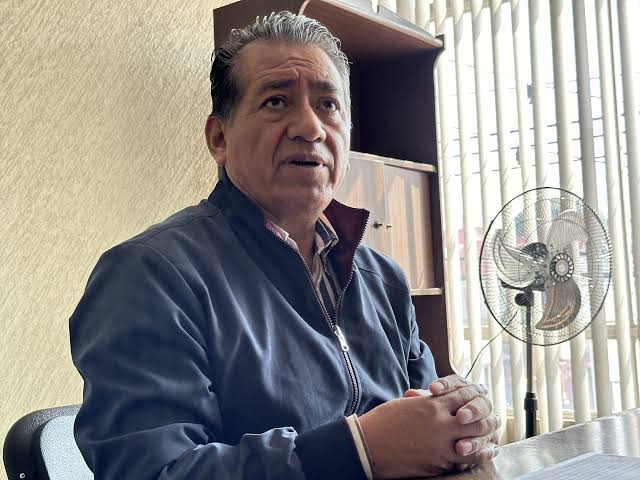 Hombre mayor sentado en una oficina, con expresión pensativa, vestido con chaqueta azul y camisa a rayas, en un ambiente con cortinas y ventilador en el fondo.