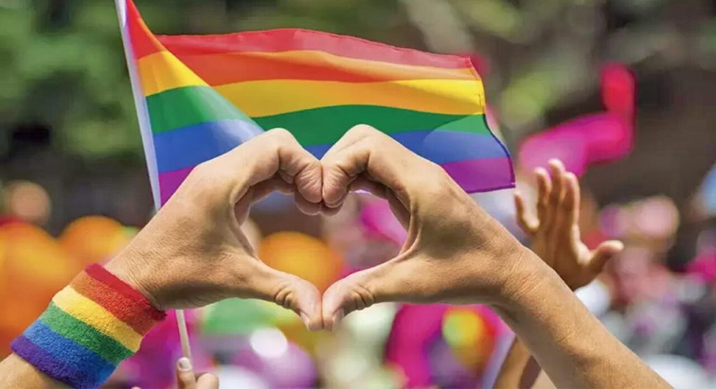 Una mano con una pulsera de arcoíris formando un corazón, en un evento del Día del Orgullo LGBTQ+ con banderas y personas en el fondo.