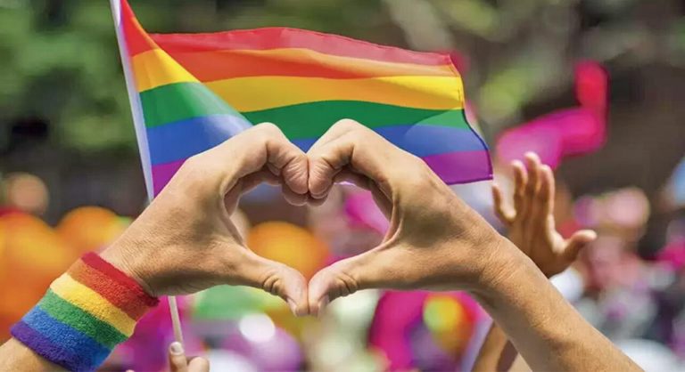 Una mano con una pulsera de arcoíris formando un corazón, en un evento del Día del Orgullo LGBTQ+ con banderas y personas en el fondo.