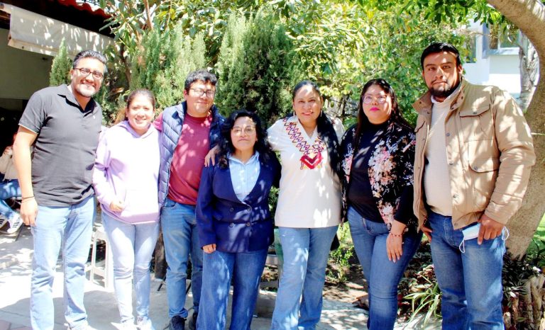 Junta de siete personas sonriendo y posando al aire libre en un entorno con árboles y plantas verdes, en un día soleado.