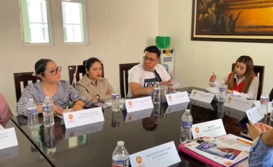 Reunión de cinco personas en una mesa de conferencia, discutiendo temas importantes en un entorno de oficina moderna. Participantes con papeles, laptops y botellas de agua en la mesa, en un ambiente f.