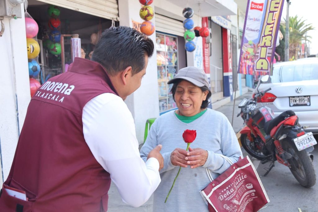 Una persona de Morena Tlaxcala entregando una rosa a una mujer en la calle, en un entorno urbano con tiendas y motocicletas visibles en el fondo.
