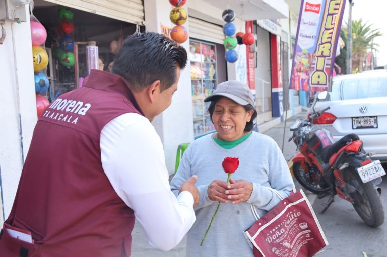 Una persona de Morena Tlaxcala entregando una rosa a una mujer en la calle, en un entorno urbano con tiendas y motocicletas visibles en el fondo.