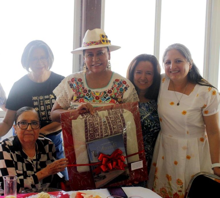 Grupo de mujeres en una celebración, una de ellas con sombrero y vestido tradicional, sosteniendo un regalo envuelto en papel decorativo.