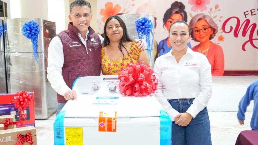 Lavadora nueva en exhibición en una tienda, con tres personas sonriendo y posando para la foto, en un evento de promoción con decoraciones coloridas y un fondo con personajes animados.