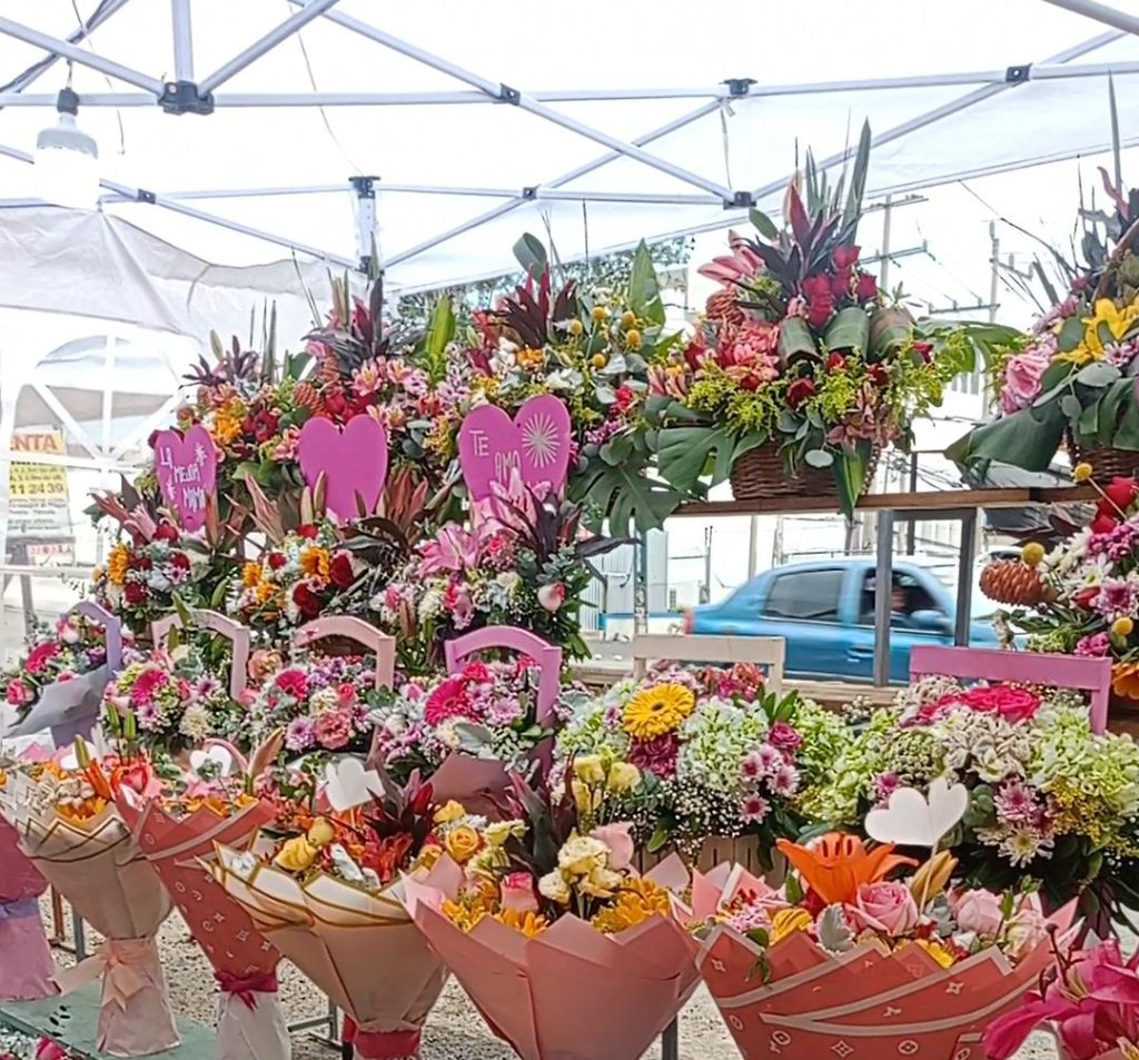 Ramo de flores variadas en un puesto de mercado, con corazones de papel y mensajes de amor, en un ambiente al aire libre con toldos y vehículos en movimiento en el fondo.
