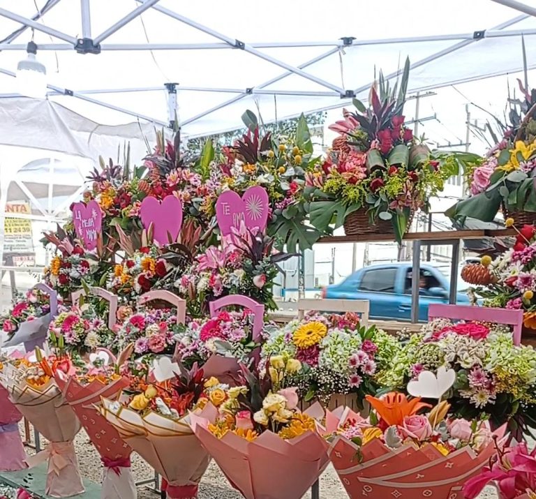 Ramo de flores variadas en un puesto de mercado, con corazones de papel y mensajes de amor, en un ambiente al aire libre con toldos y vehículos en movimiento en el fondo.