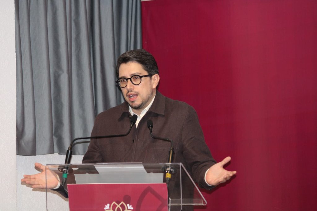 Hombre hablando en conferencia con fondo gris y rojo, usando gafas y chaqueta oscura, en un evento formal.