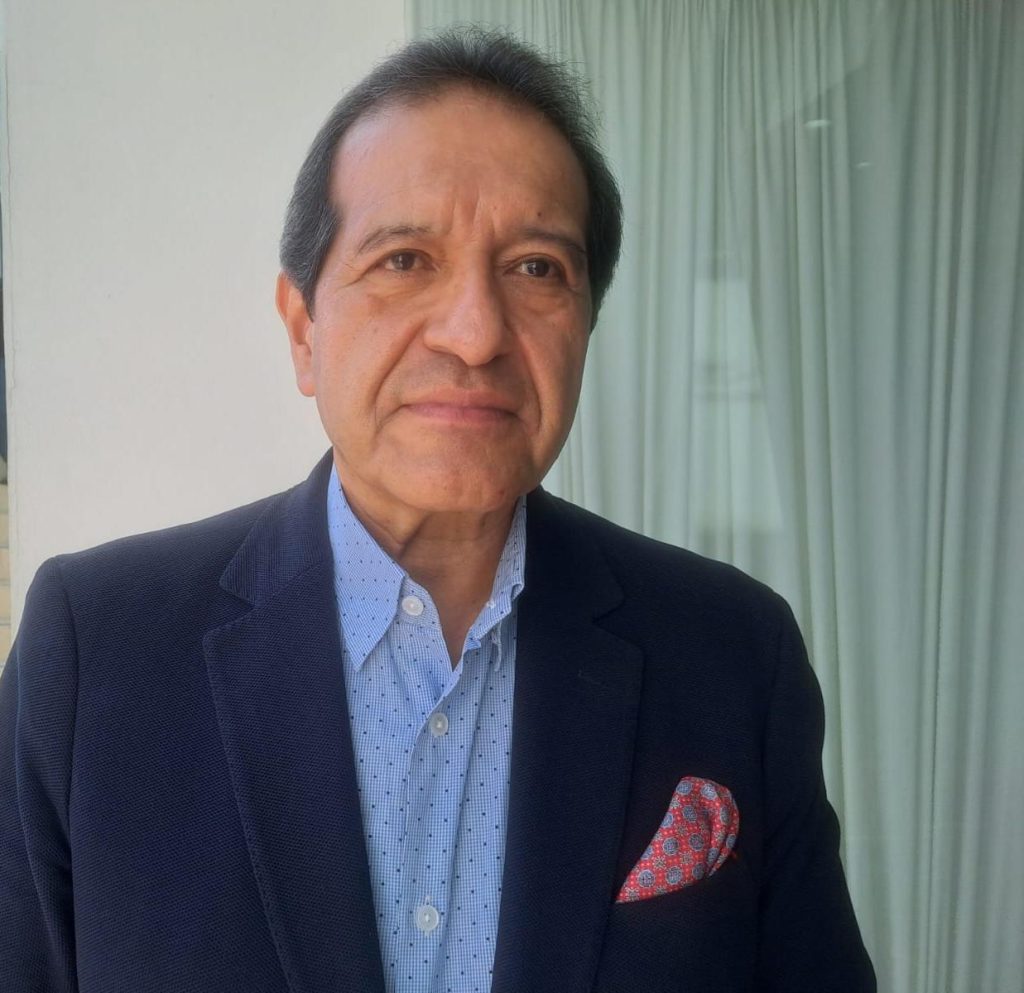 Retrato de un hombre con traje azul y camisa de vestir, en un entorno interior con cortinas blancas y luz natural.