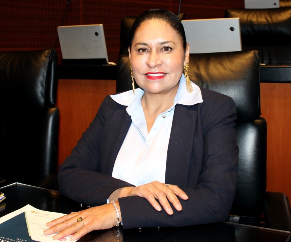 Mujer de negocios en una reunión formal, vestida con traje oscuro y blusa blanca, en un entorno de oficina moderna con computadoras y mobiliario ejecutivo.