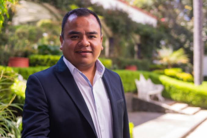 Hombre con traje oscuro y camisa blanca en un parque con vegetación y bancos, sonriendo para una foto profesional.