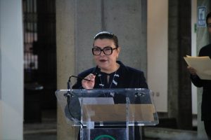 Mujer hablando en un podio durante una conferencia o evento oficial en un espacio público, con fondo de columnas y personas observando.