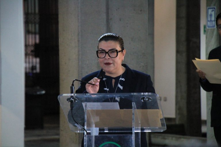 Mujer hablando en un podio durante una conferencia o evento oficial en un espacio público, con fondo de columnas y personas observando.