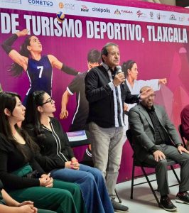 Hombre hablando en evento deportivo en Tlaxcala, con mujeres y un fondo rosa con texto sobre turismo deportivo.