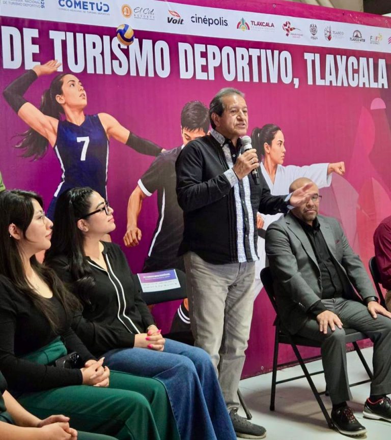 Hombre hablando en evento deportivo en Tlaxcala, con mujeres y un fondo rosa con texto sobre turismo deportivo.