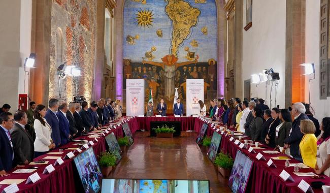 Reunión oficial en un salón con mural del mundo y participantes de pie en una ceremonia formal.