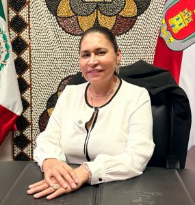 Una mujer sentada en una oficina con banderas de México y Tlaxcala al fondo, luciendo una blusa blanca y una expresión amigable.